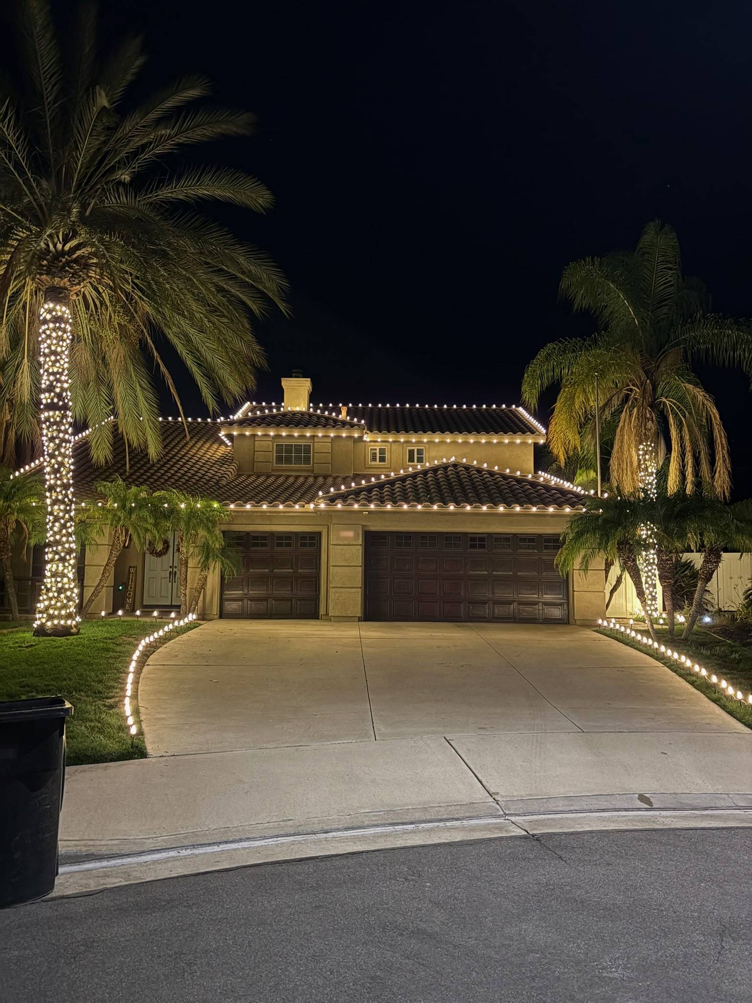 Warm-white C9 roofline on both stories of a Spanish-tile home with two lit palm trunk wraps and driveway edge markers